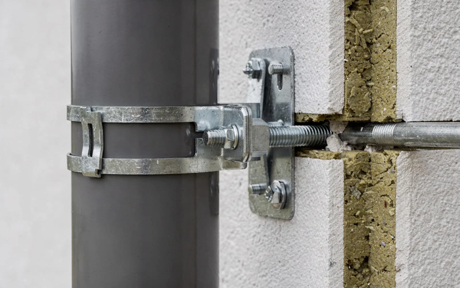 Verzinkte Wandkonsole hält das Rohr vor einer verputzten Wand; ein Gewindestab führt durch die Dämmschicht ins Mauerwerk dahinter.