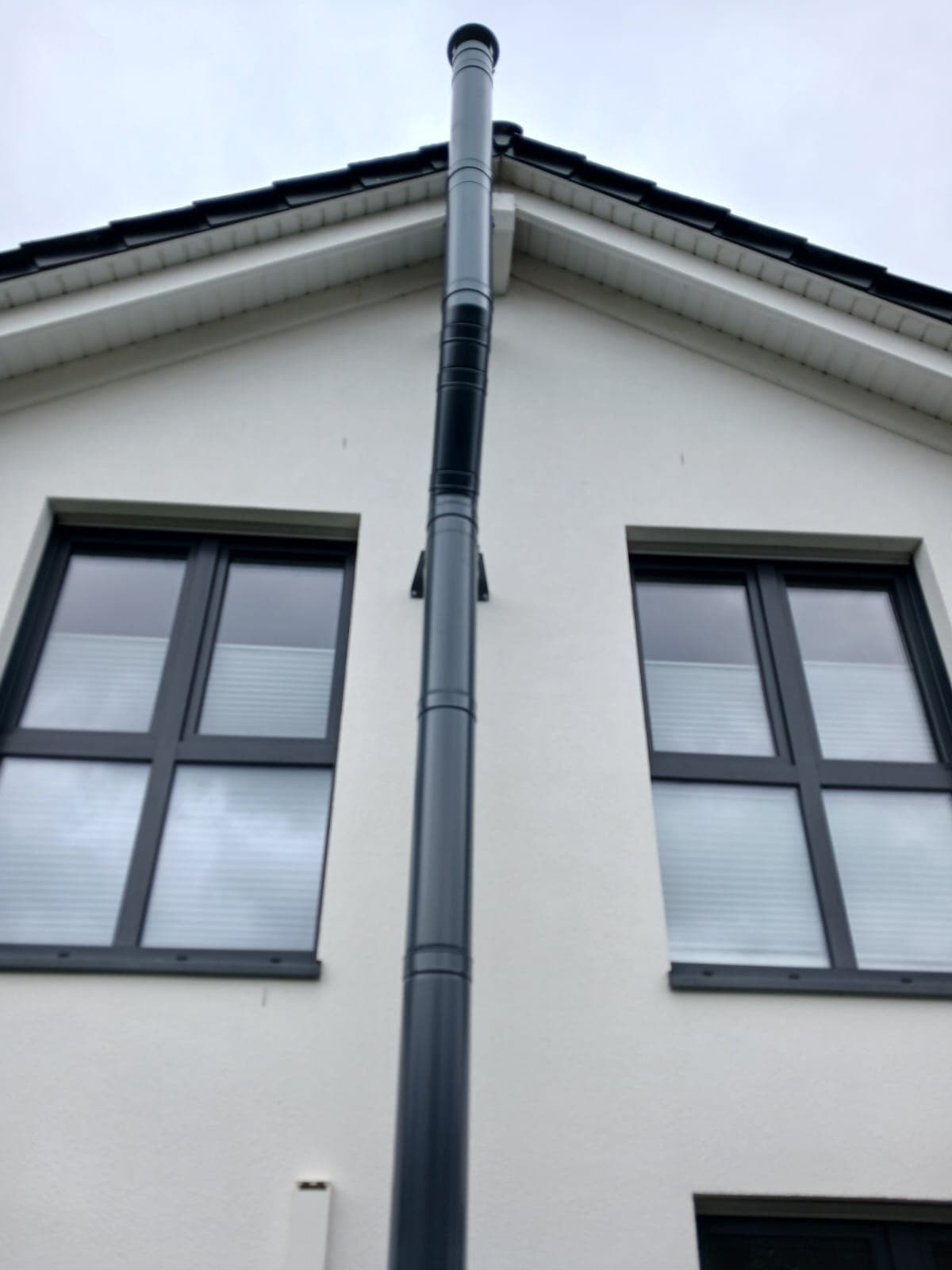 Doppelwandiger Edelstahlschornstein in Anthrazit an der weißen Giebelwand eines Einfamilienhauses, senkrecht zwischen zwei Fenstern geführt, die Mündung überragt den First.
